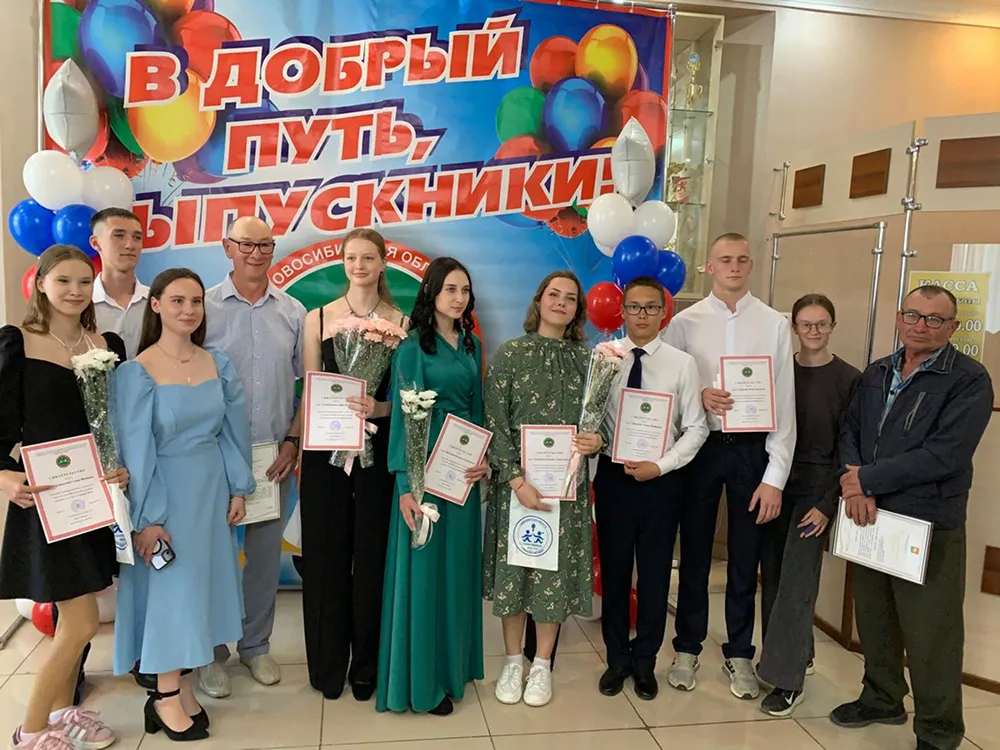 Выпускники с дипломами и цветами на торжественной церемонии, групповое фото на фоне баннера «В добрый путь, выпускники!»